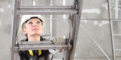 A woman wearing a hard hat, looking up and climbing a ladder.